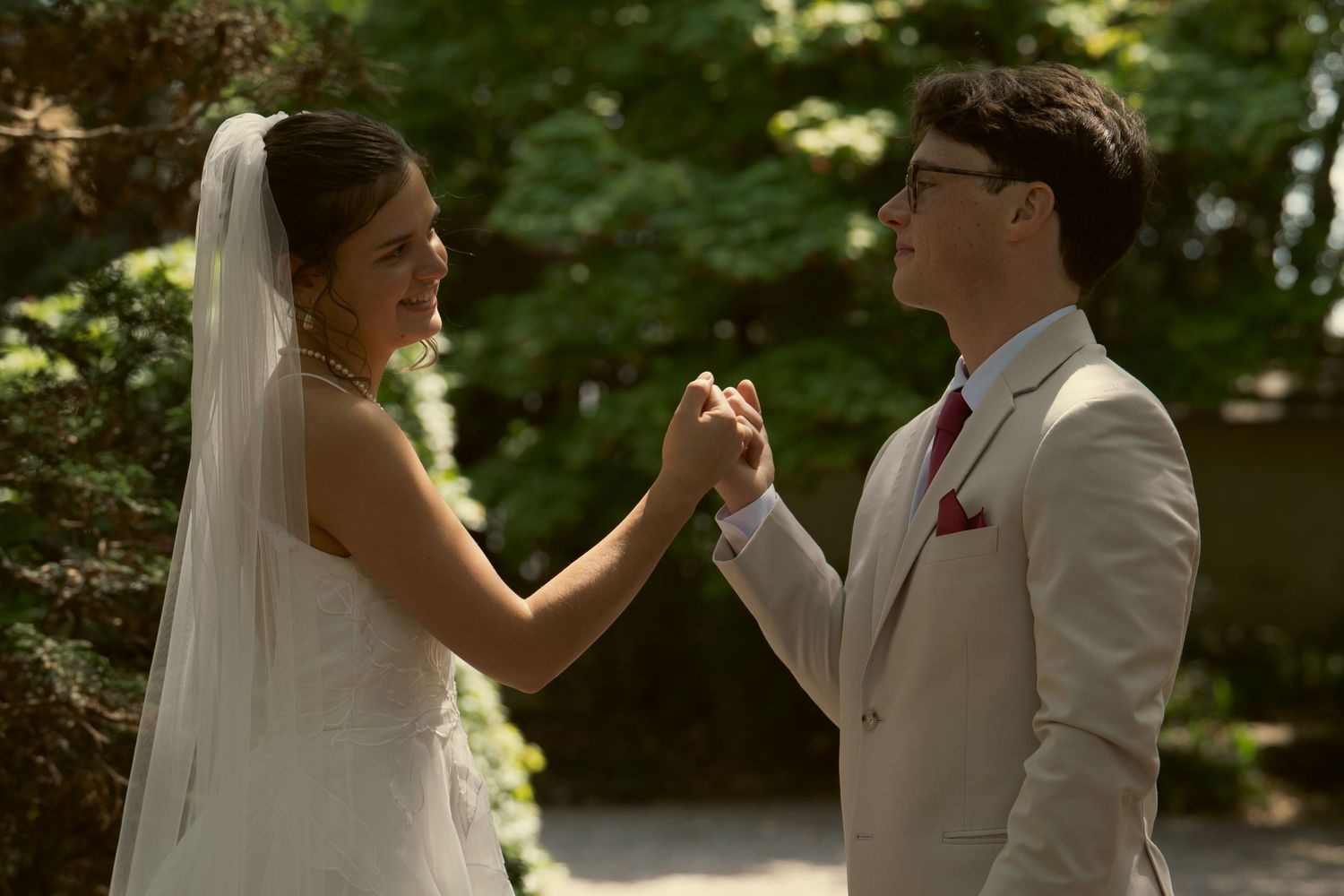 Bride and groom hold hands and smile at each other outdoors in a sunlit garden setting.