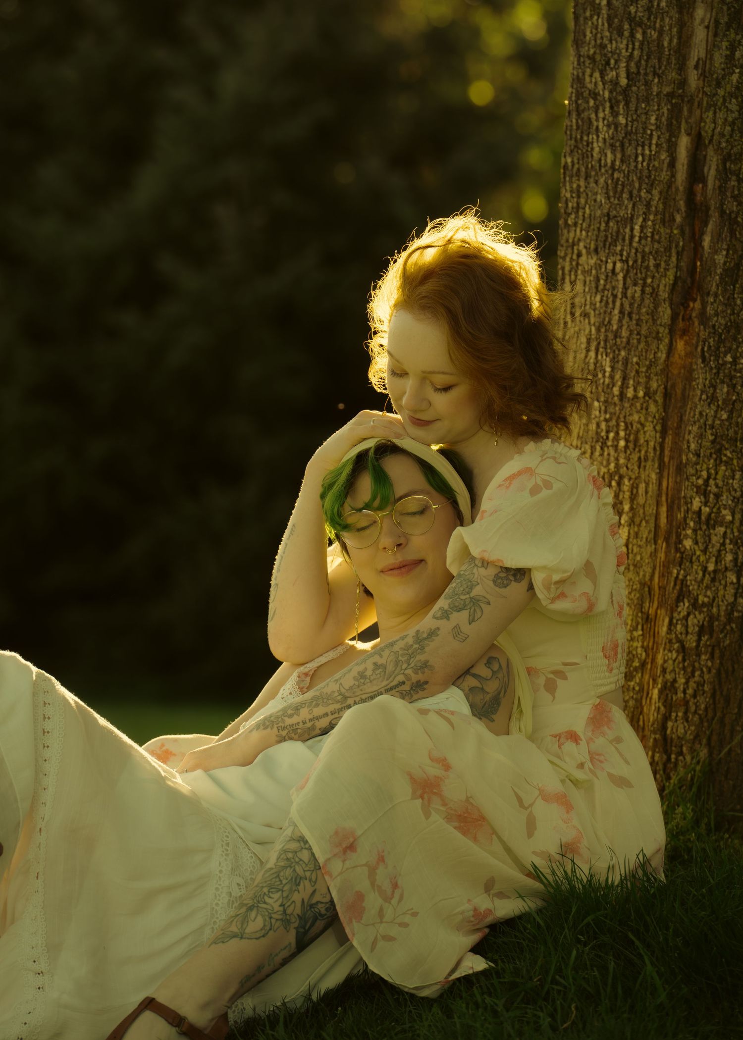Two women in floral dresses share a tender moment leaning against a tree in golden sunlight.