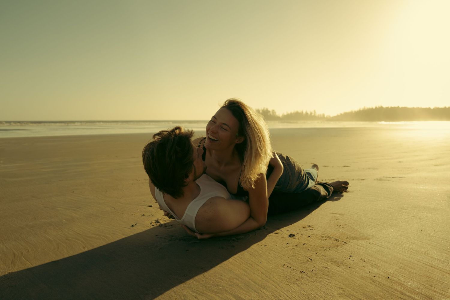 Romantic couple lying on a sandy beach at sunset, sharing an intimate moment with golden light.