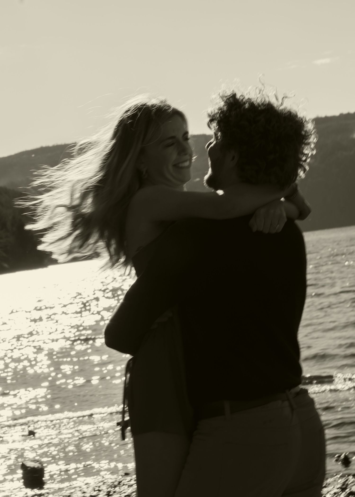 Black and white photo of a couple embracing and smiling by a sunlit lake shore with hills in the background.