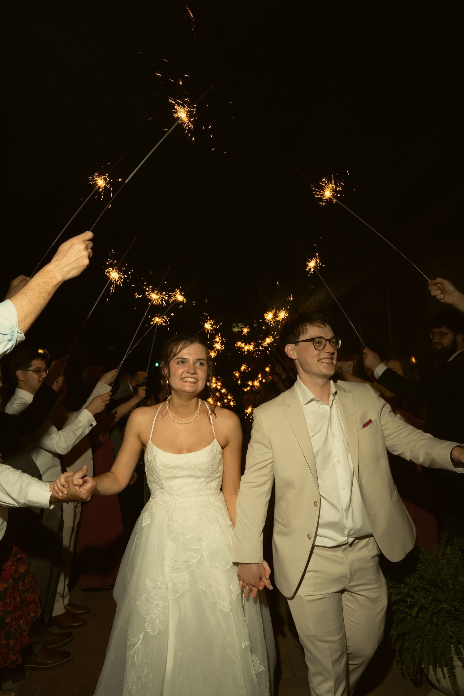 Bride and groom exit wedding reception through a glowing sparkler tunnel at night surrounded by guests.