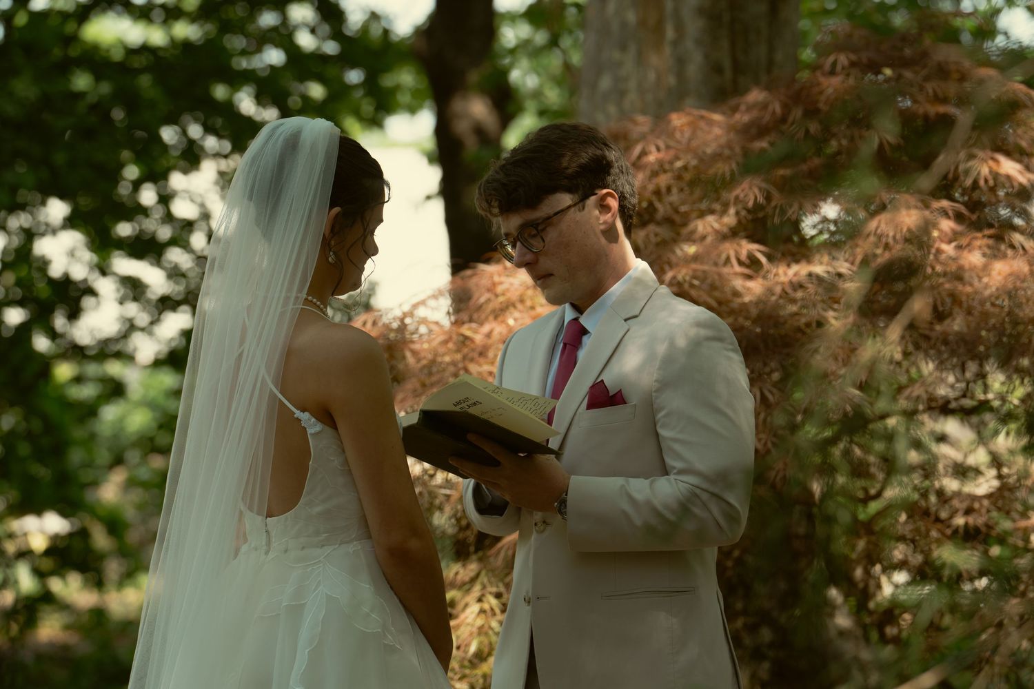 Groom in tan suit reads vows to veiled bride during outdoor forest wedding ceremony.