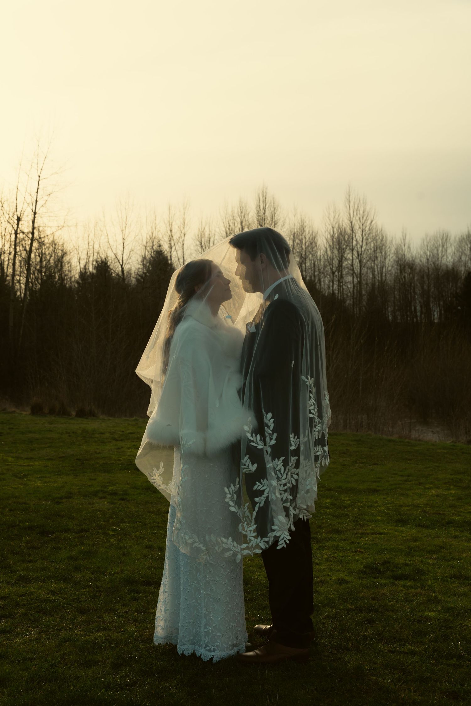 Two figures in ethereal white dresses stand silhouetted against a moody sunset landscape in a field.