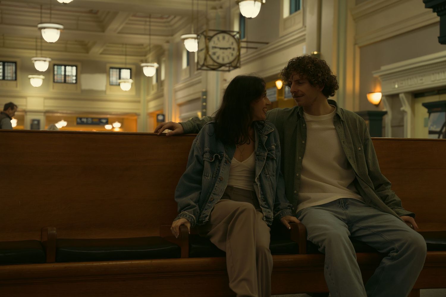 A cozy indoor scene shows two figures conversing in a warmly lit institutional space.