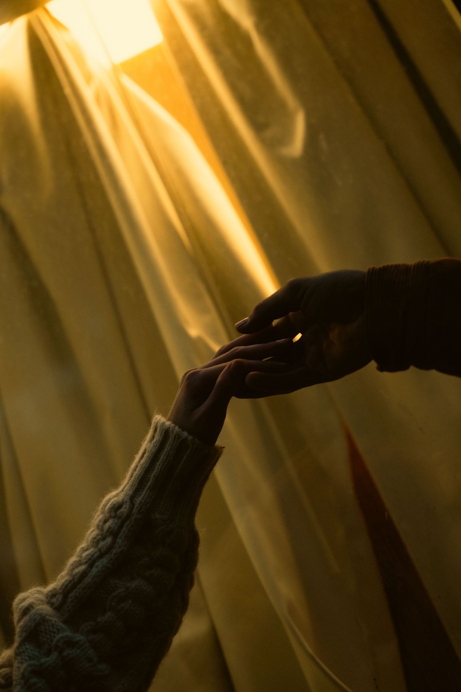 Two hands reaching toward each other against golden curtains in dramatic lighting.