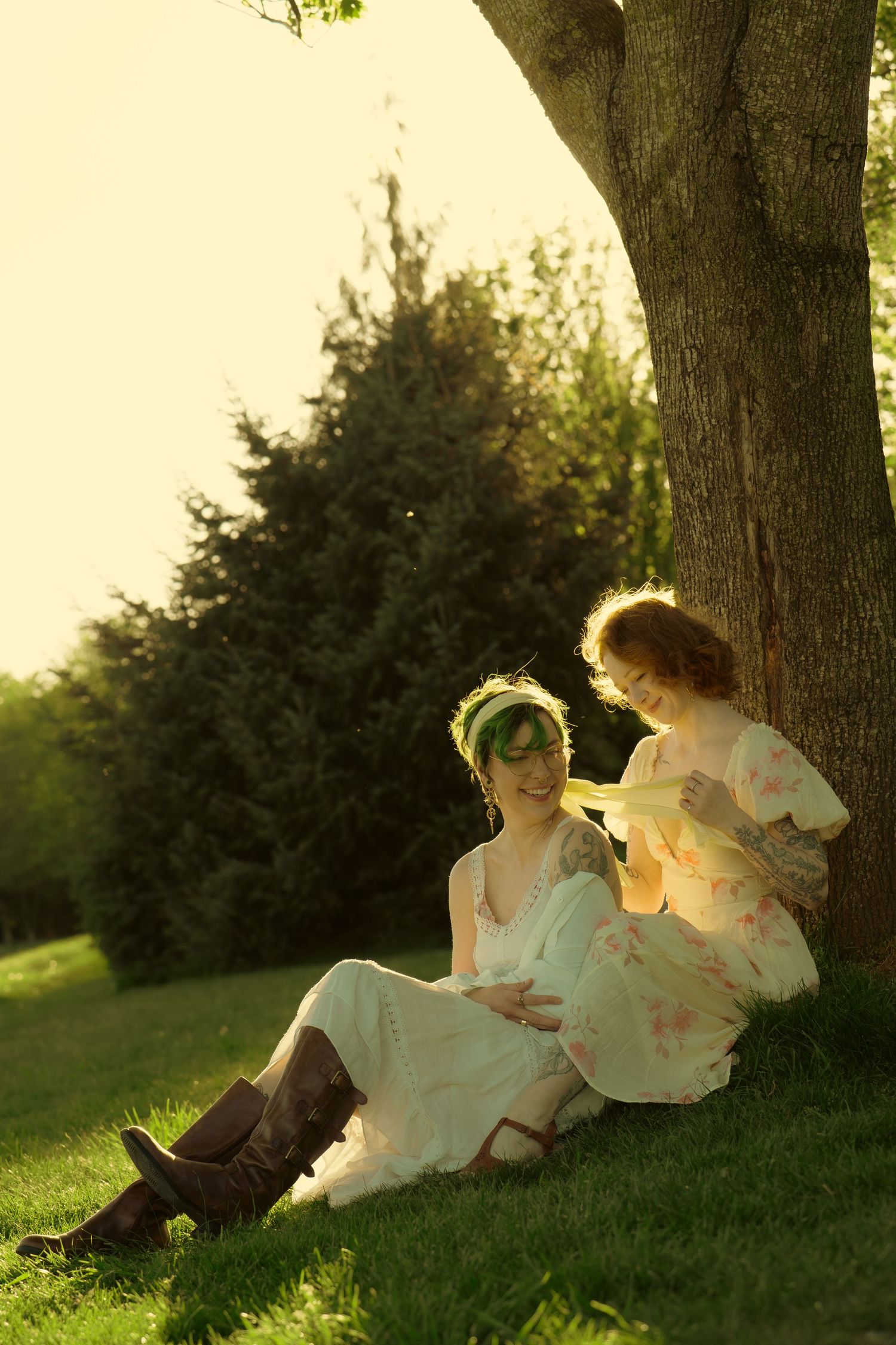 Two women in vintage dresses sit under a large tree in a sunlit park, sharing a quiet moment.