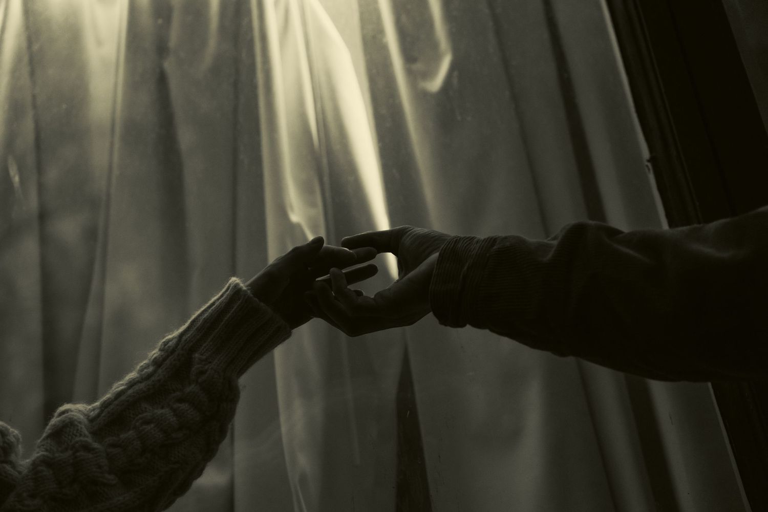 Two hands meeting in silhouette against white curtains in monochrome.