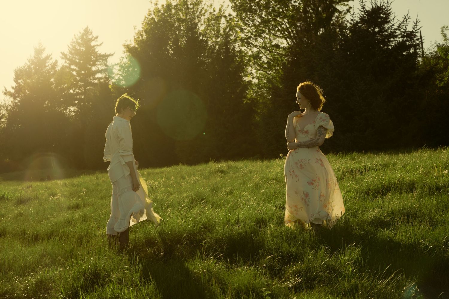 Two women in flowing white dresses walk through a sunlit green meadow surrounded by trees at golden hour.