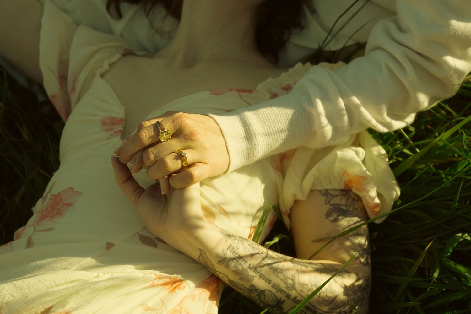 Woman in floral dress lying on grass with clasped hands wearing gold rings, tattoo visible on wrist.