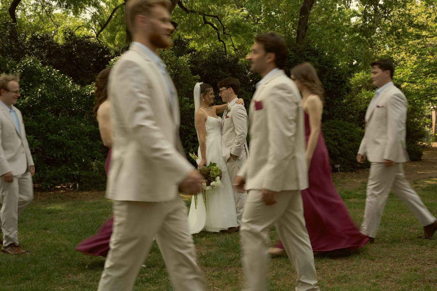 Wedding party walking outdoors in formal attire, groomsmen in tan suits and bridesmaids in burgundy dresses.