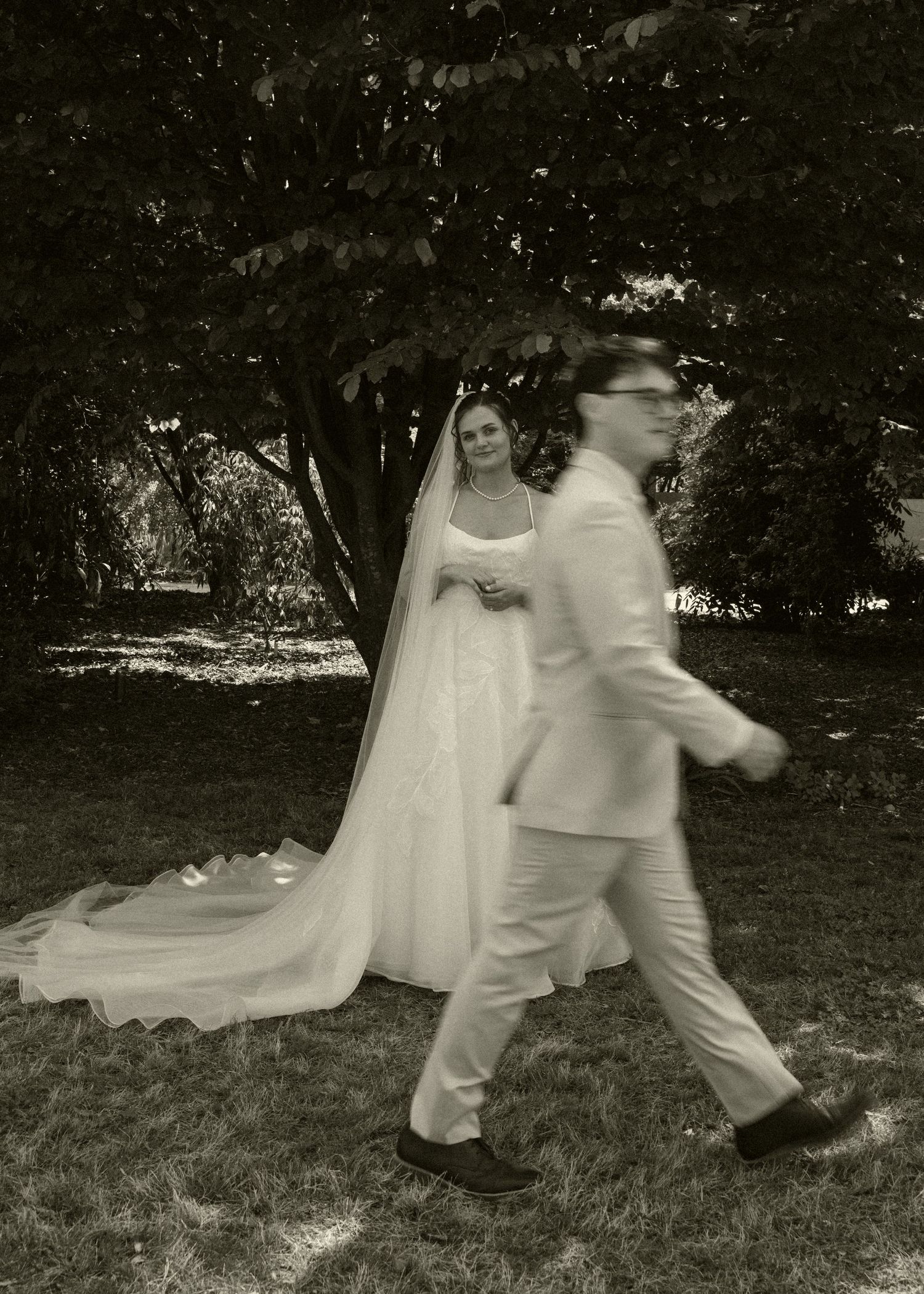 Black and white photo of bride in veil watching groom walk past under large tree.