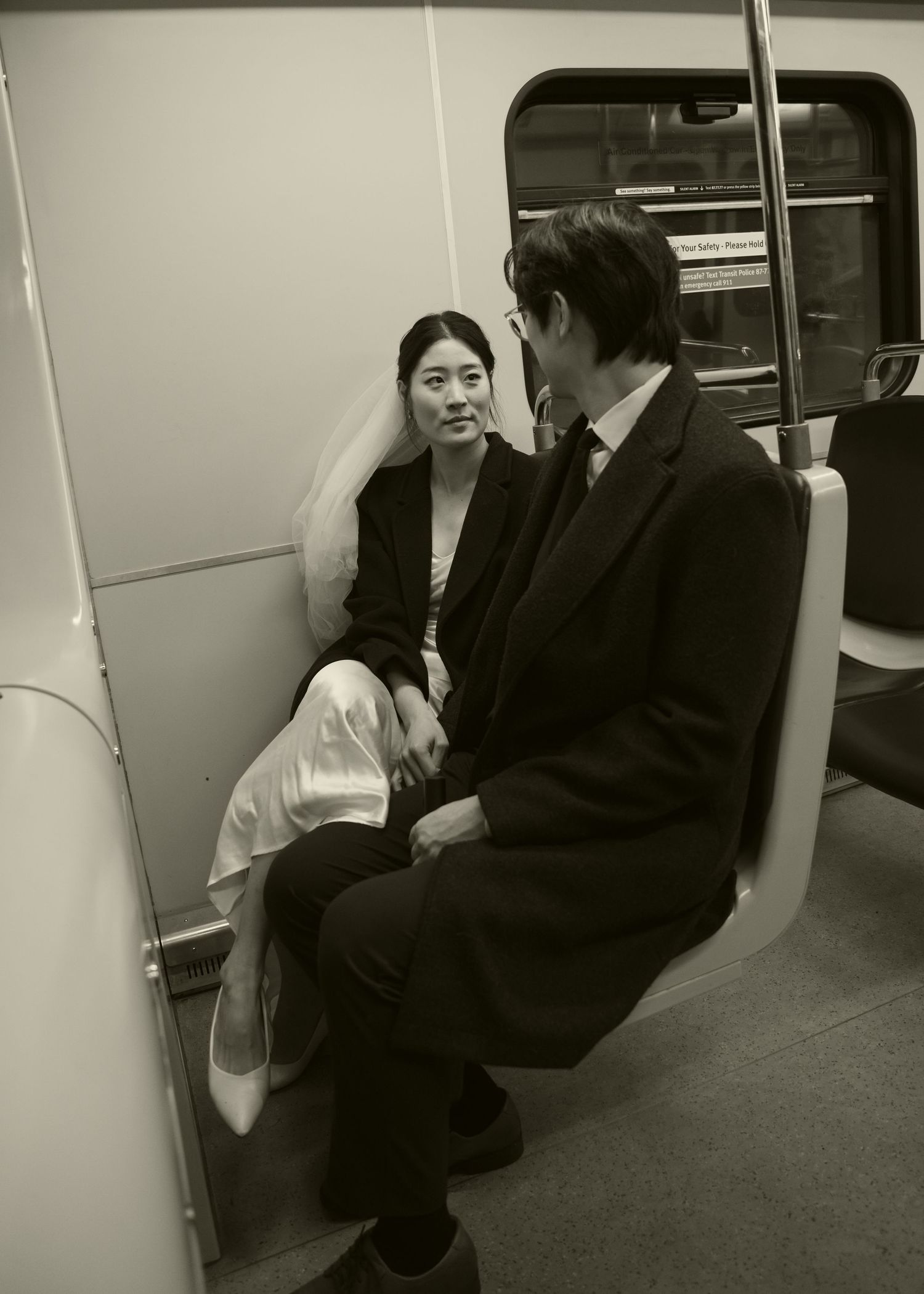 Black and white photo of a bride and groom in formal attire sitting together on a subway train.
