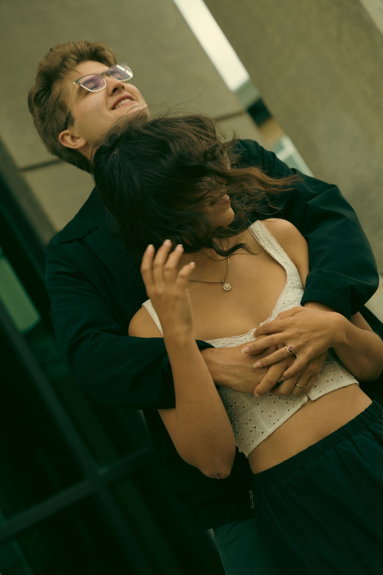 A man in dark jacket embraces a woman with flowing hair in a white bralette top in an urban setting.