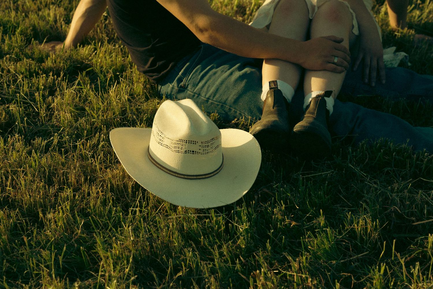 White cowboy hat resting on green grass beside a person sitting outdoors in warm golden light.