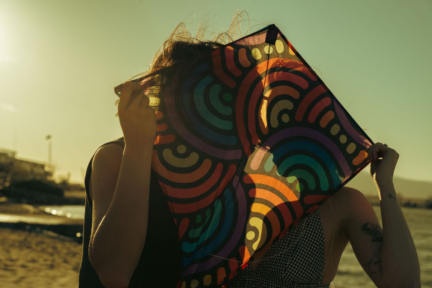 Woman holding colorful spiral patterned kite in front of her face on a sunny beach at sunset.