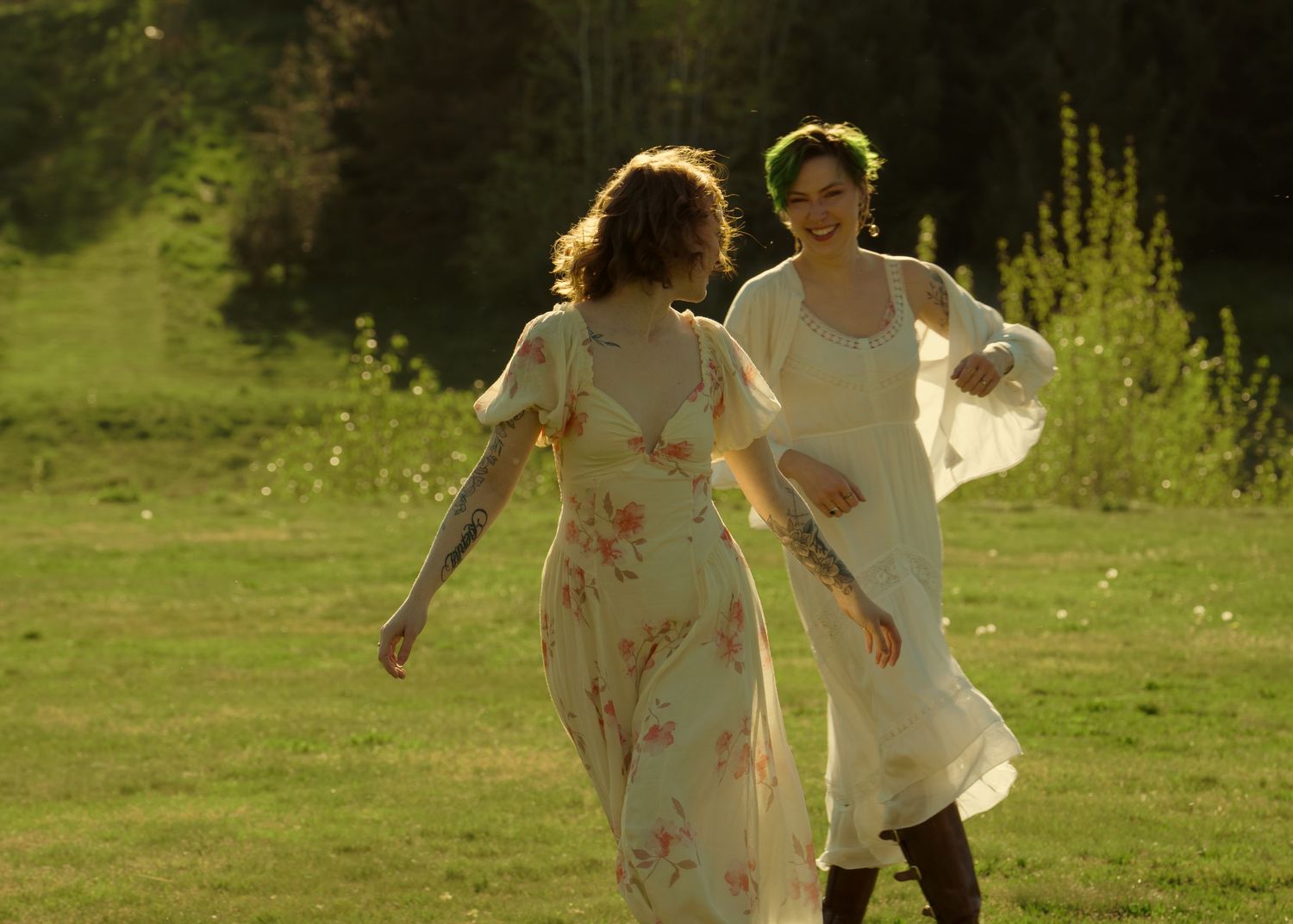 Two women in vintage floral dresses walking together across a sunlit green meadow at golden hour.