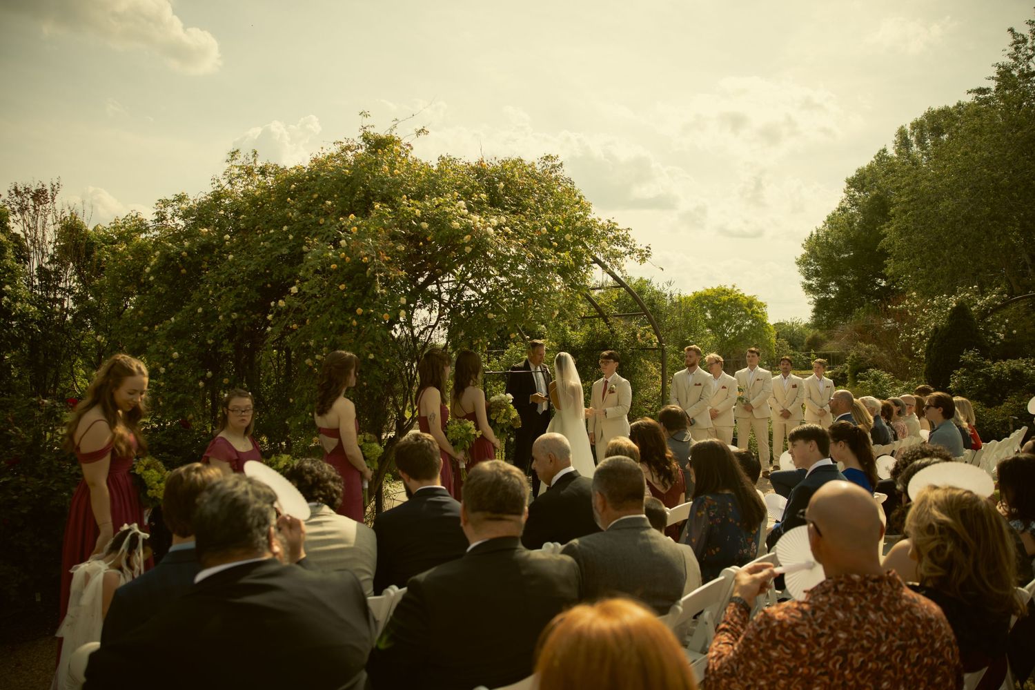Outdoor wedding ceremony in a garden with guests seated, bridal party in burgundy dresses standing near a floral arch.