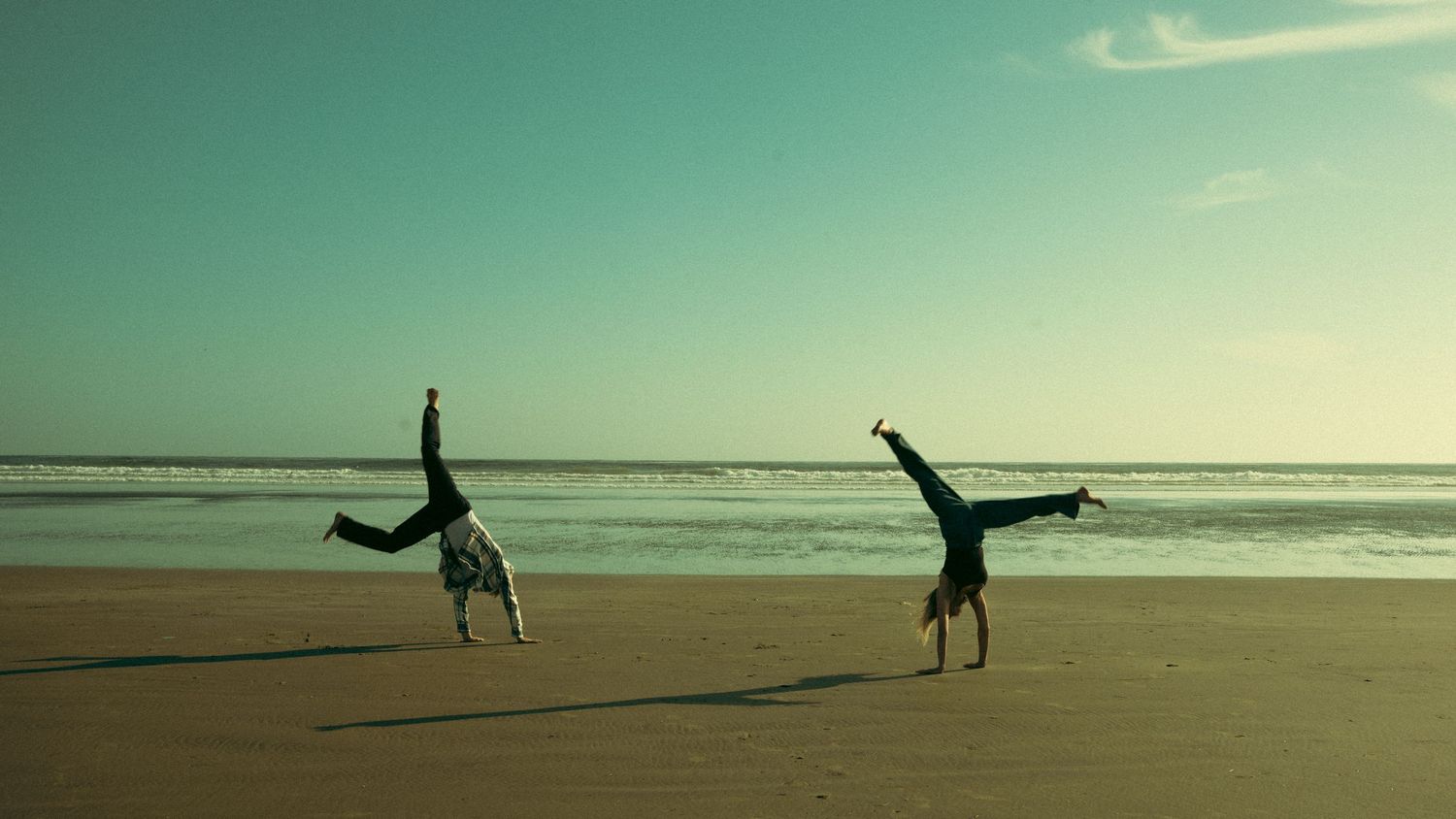 Two people doing cartwheels on beach against turquoise sky.