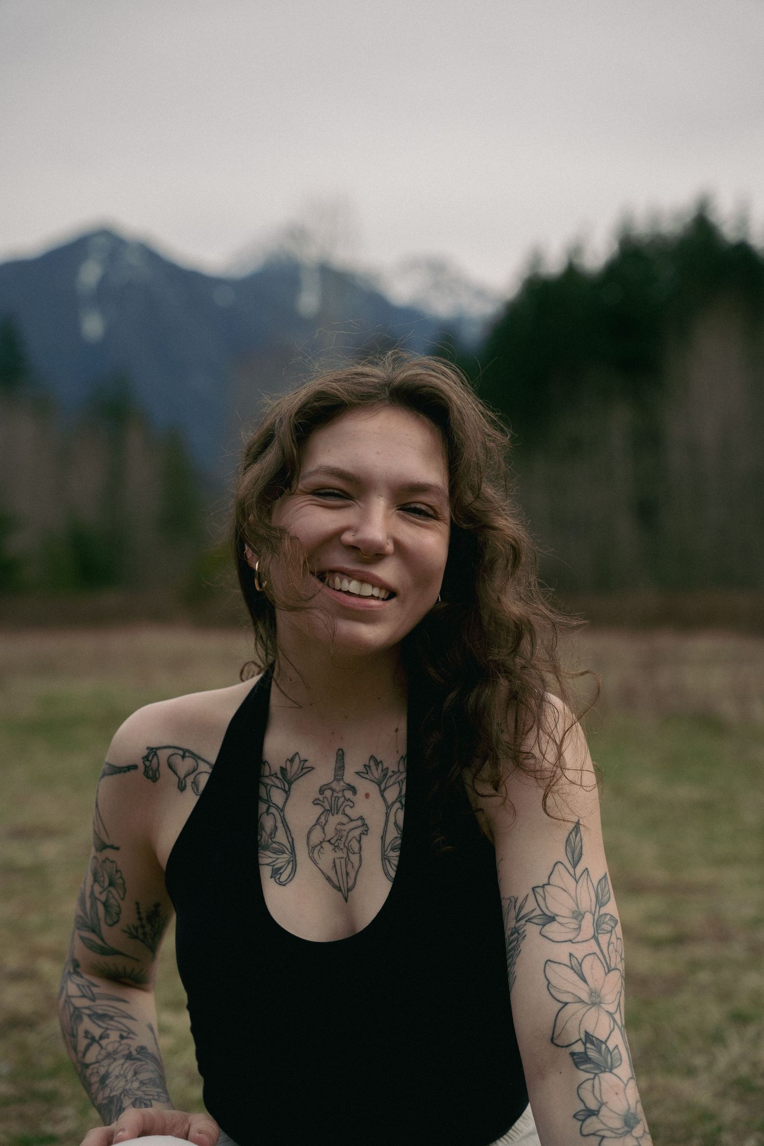 Person in black tank top with artistic tattoos posing in front of mountain landscape.