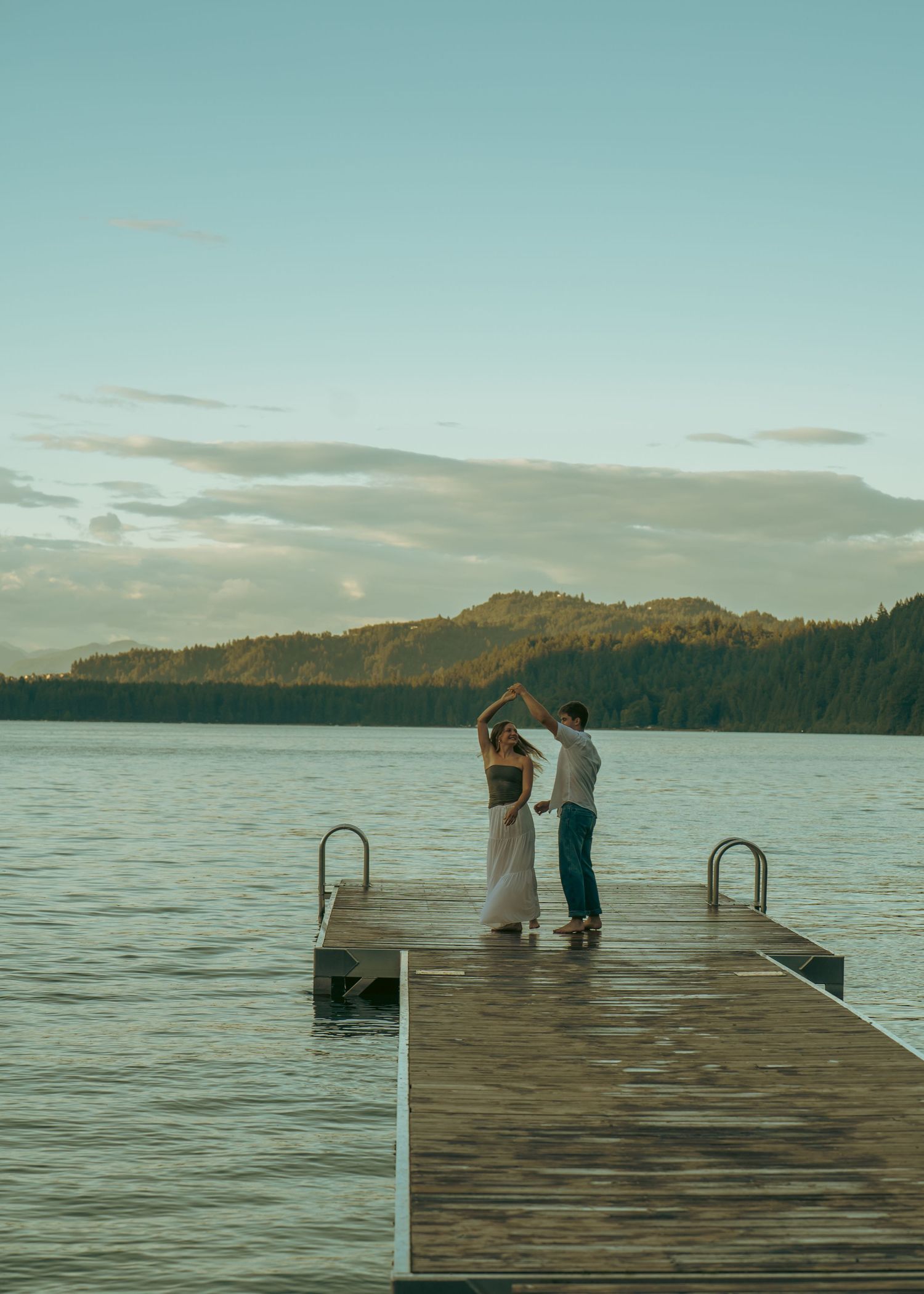A romantic couple stands at the end of a wooden dock overlooking a serene lake surrounded by mountains at sunset.
