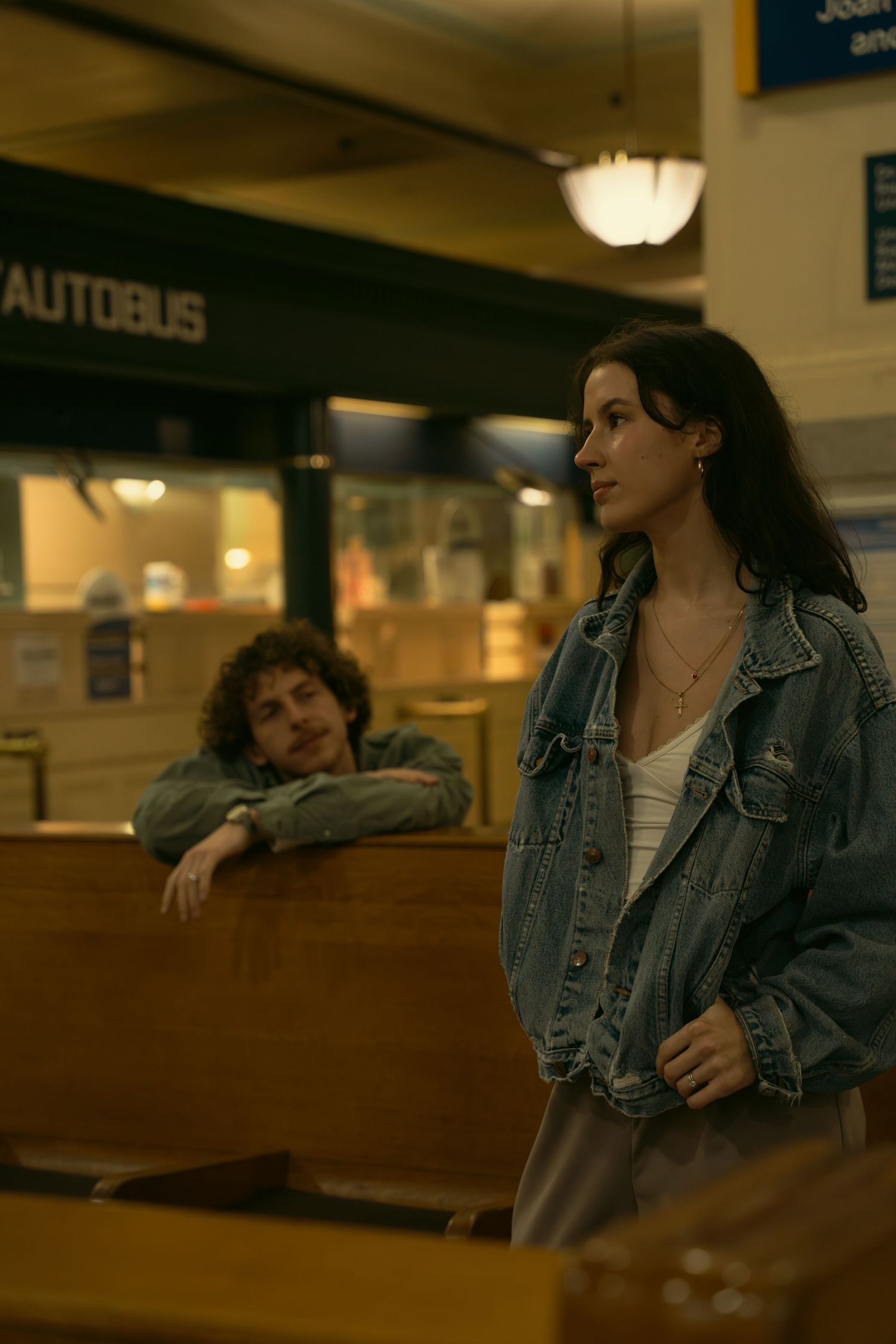 People in denim jackets converse near an 'Autobus' sign in a public building.