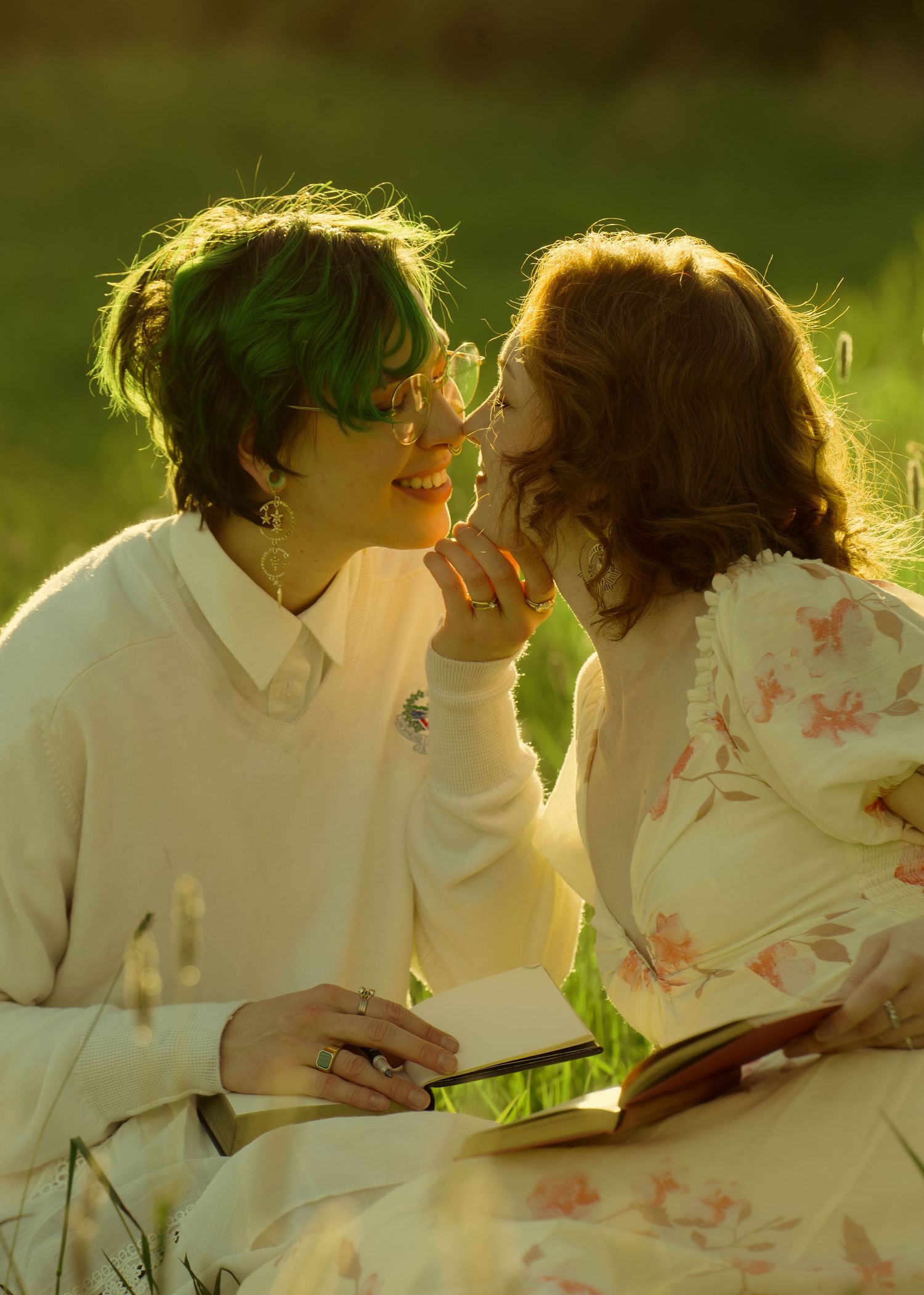 Two women share an intimate moment outdoors, one with green hair holding a book, bathed in warm golden sunlight.