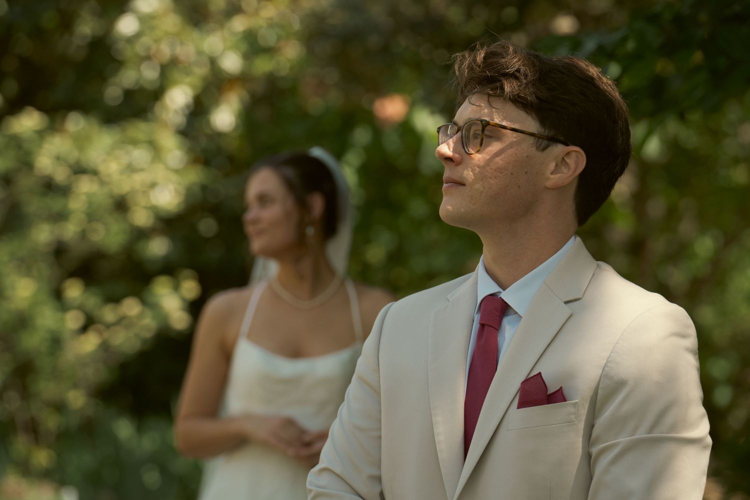 Groom in beige suit and red tie stands outdoors with bride in white dress behind him.