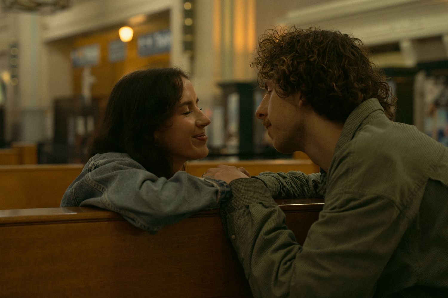 Two people in denim jackets sit close together in a dimly lit room with warm lighting.