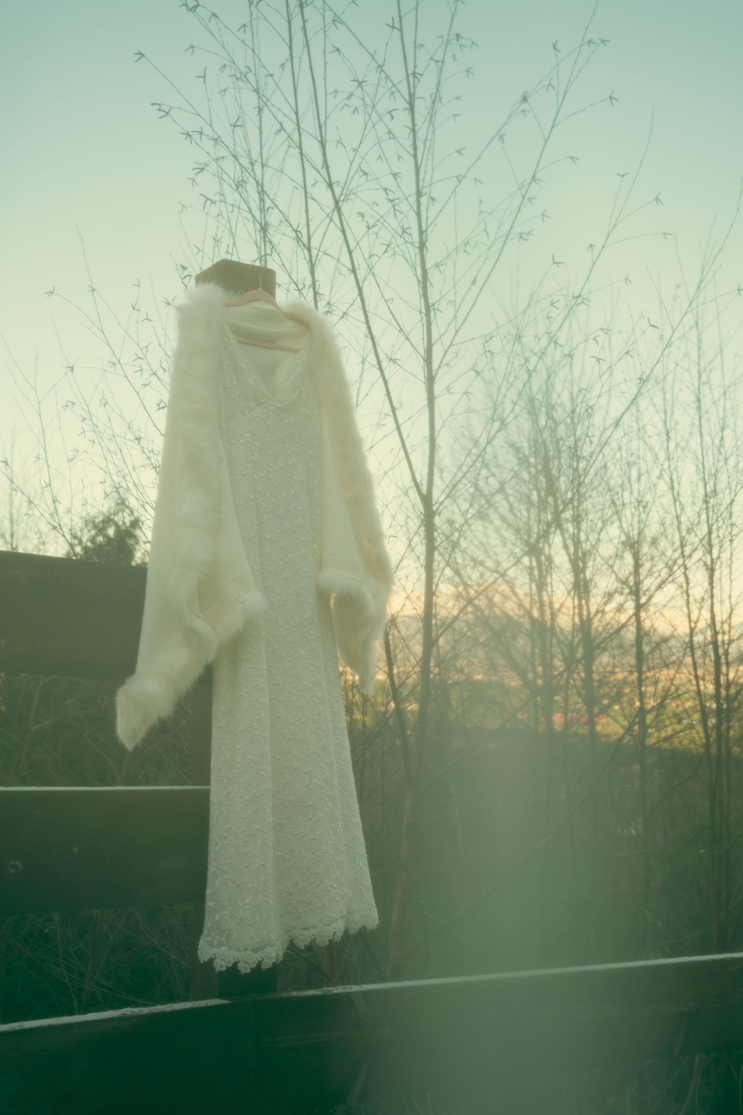 White lace wedding dress hanging outdoors against a sunset sky with bare winter trees in the background.