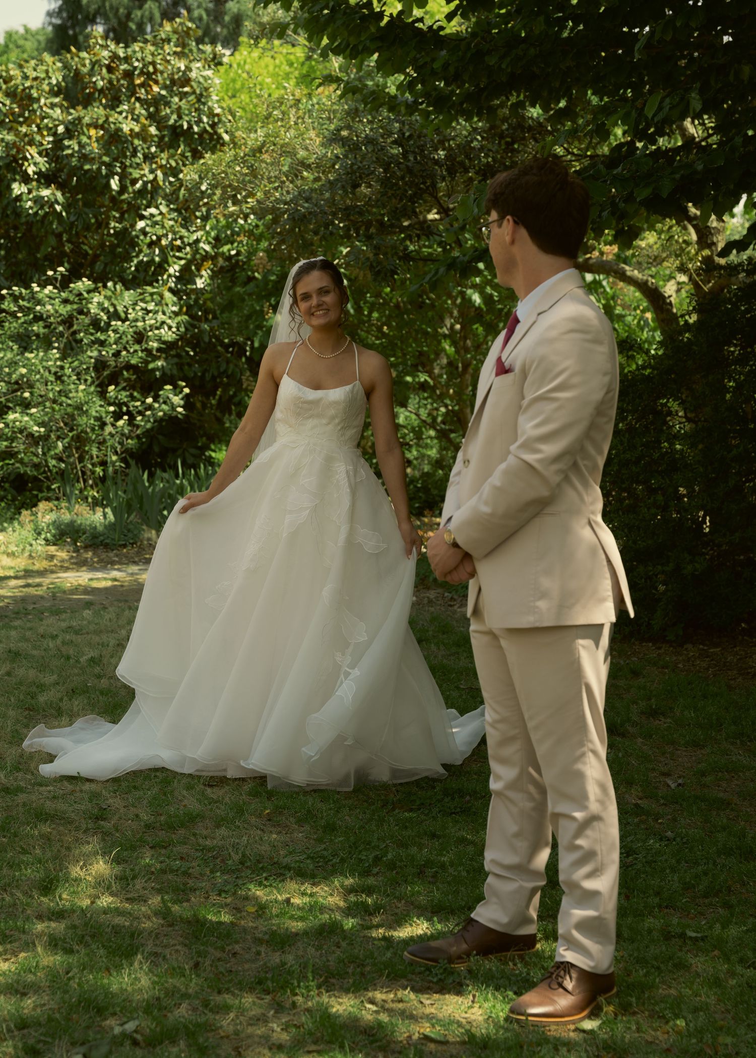 Bride in white gown and groom in tan suit hold hands outdoors in a lush garden during their wedding.