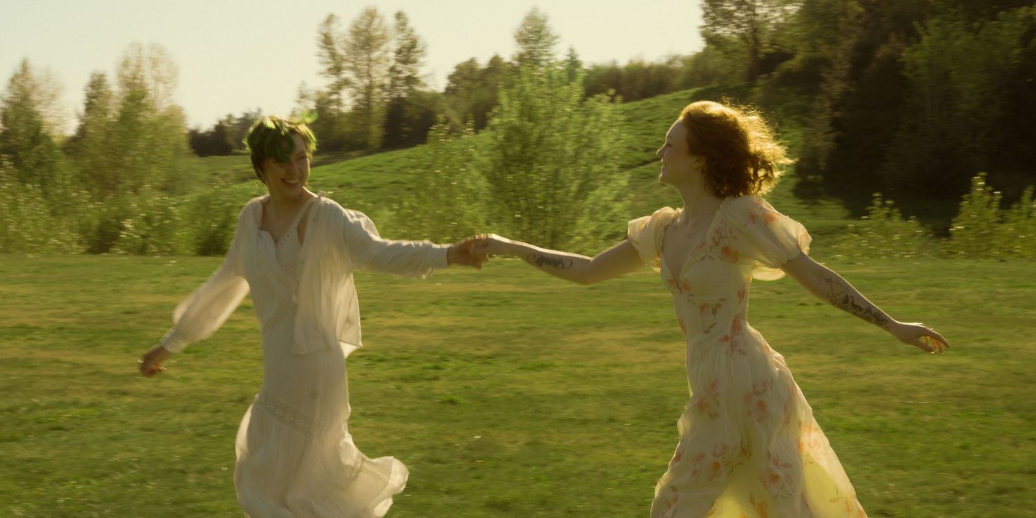 Two women in white dresses dance joyfully in a sunlit green meadow surrounded by trees.