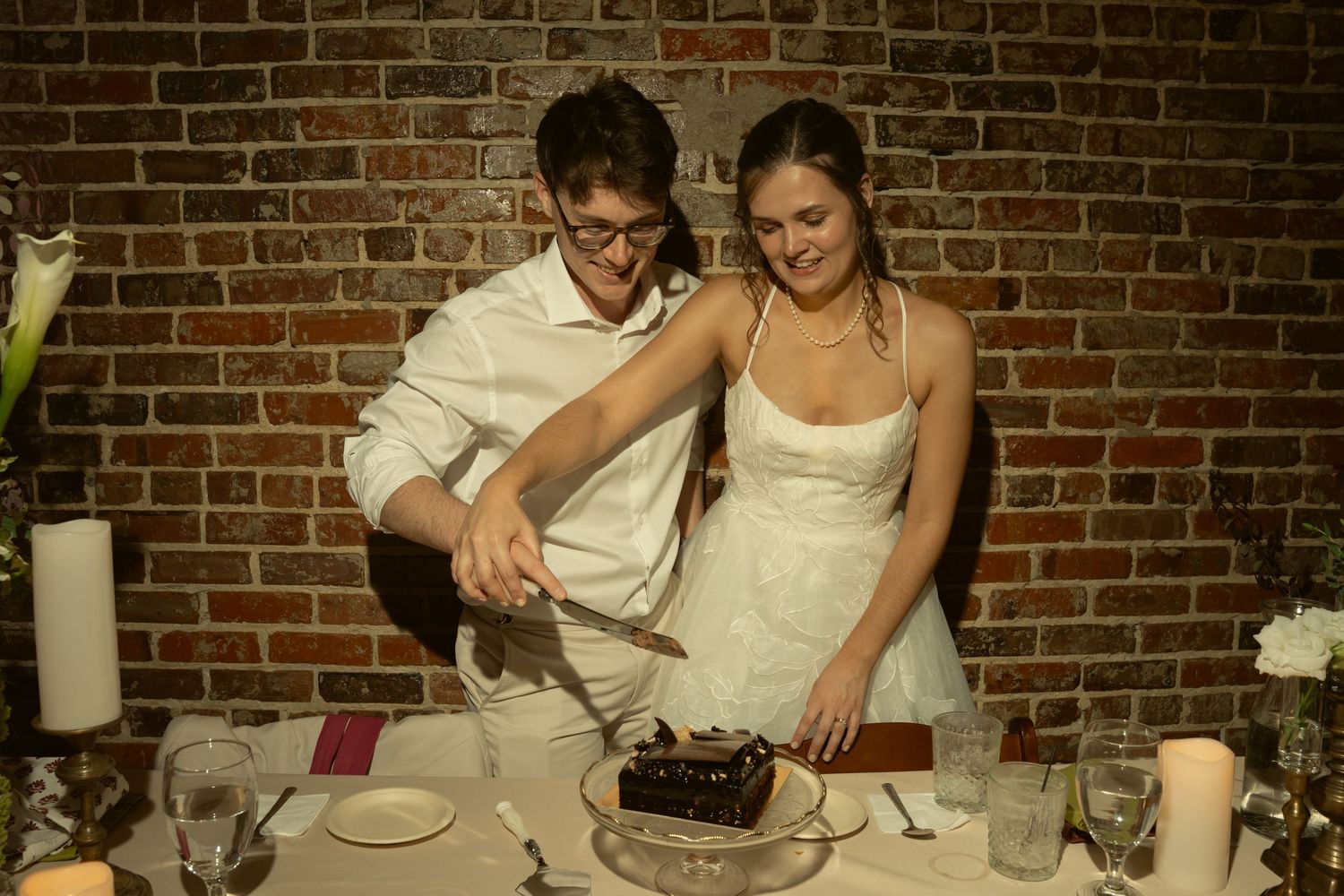 Newlyweds smiling while cutting their chocolate wedding cake at a rustic brick wall venue.
