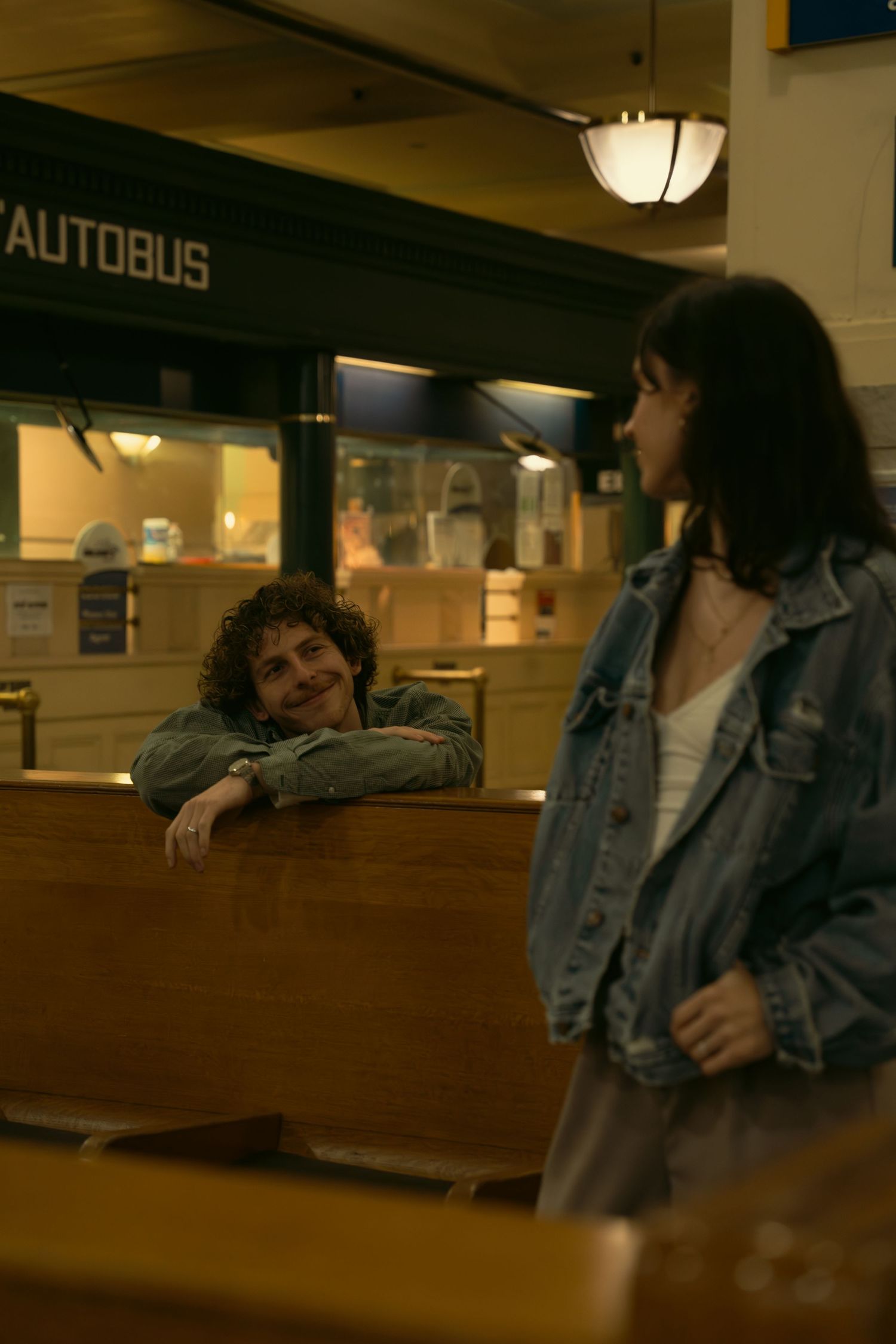 Two figures in casual attire interact in a public space with 'Autobus' signage visible.