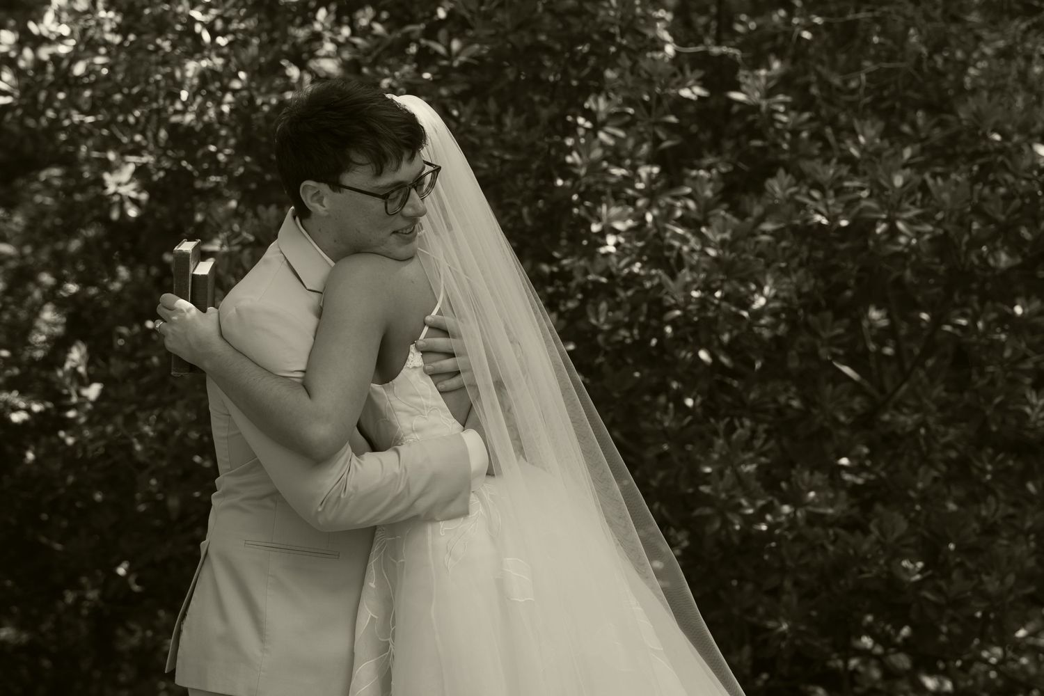 Black and white photo of newlyweds embracing tenderly outdoors, bride in white gown and veil, groom in tan suit.