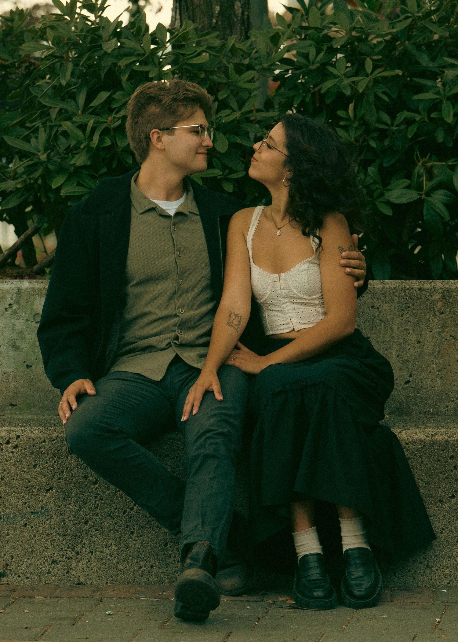 A couple sits together on a stone ledge surrounded by lush green foliage in a romantic outdoor setting.