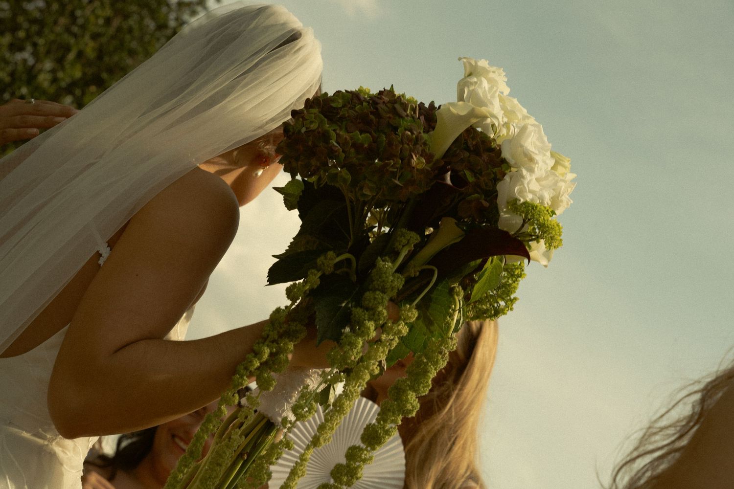Bride smelling a lush green and white wedding bouquet outdoors against a bright sky.