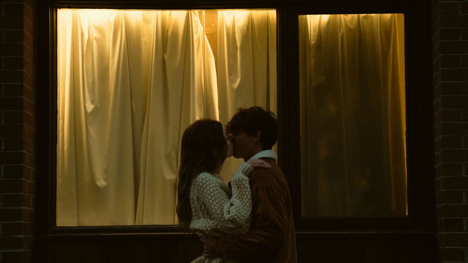 Romantic couple sharing a tender kiss silhouetted against a warmly lit window with golden curtains at night.
