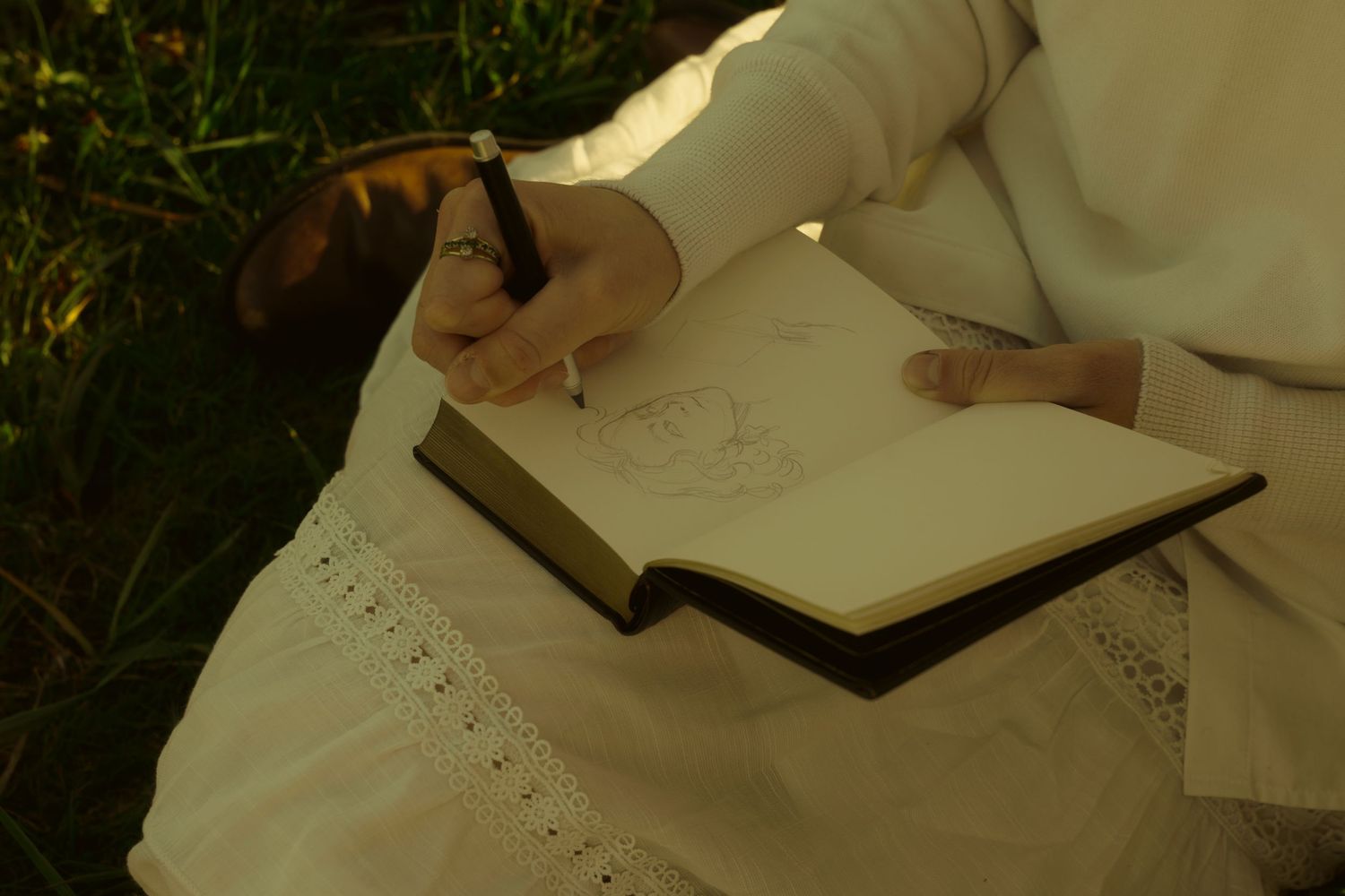 Person in white dress sketching in a journal outdoors, sitting on grass with a pen in hand.