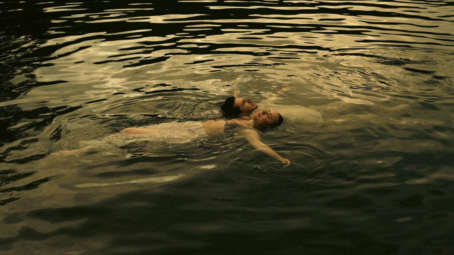 Person floating peacefully on their back in dark, calm water with golden reflections on the surface.