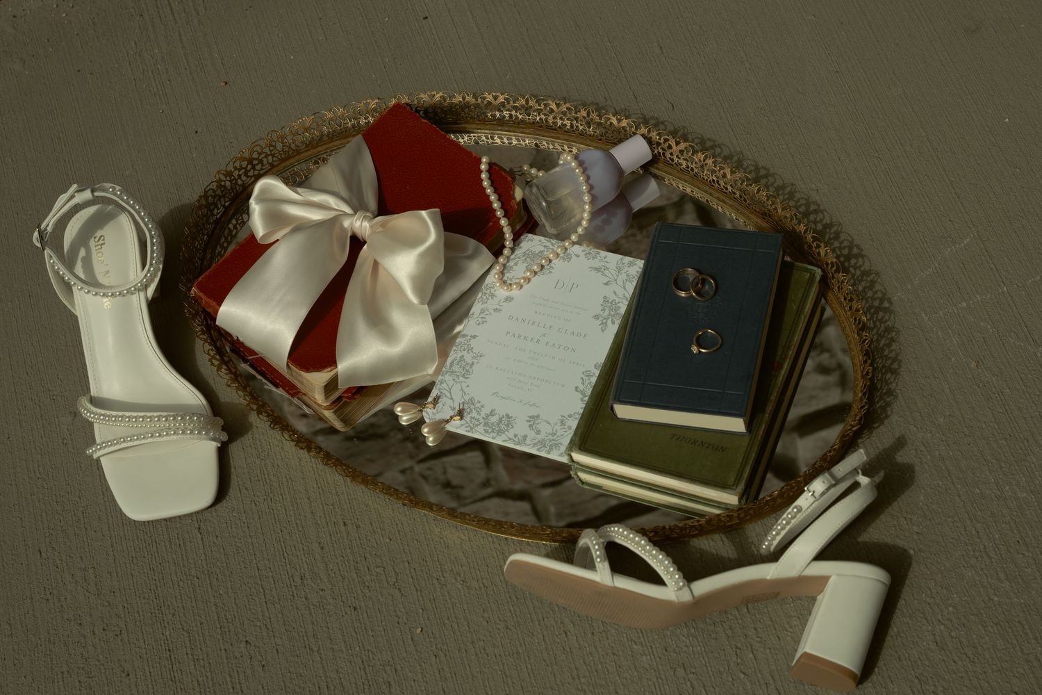 Flat lay of wedding details including white heels, pearl necklace, rings, invitation, and ribbon bow.