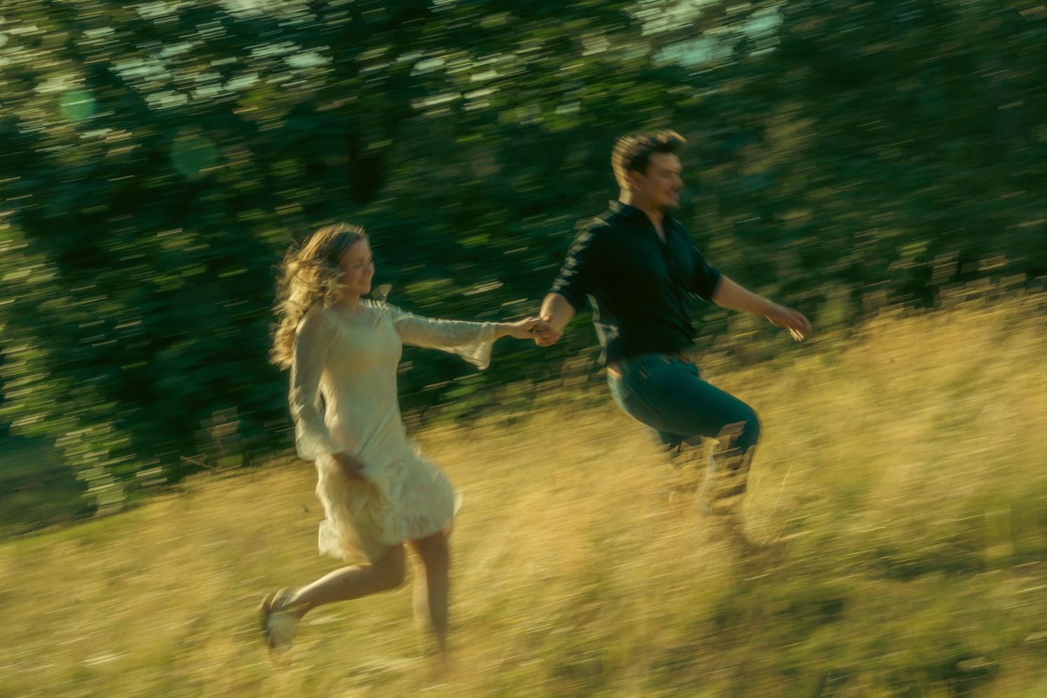 A young couple runs hand-in-hand through a sunlit green field, motion blur capturing their joyful sprint.