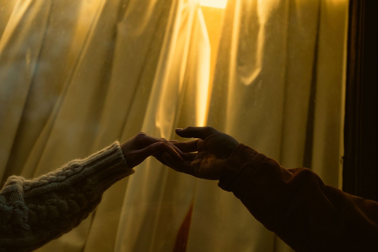 Hands reaching to touch in silhouette against sunlit golden curtains.