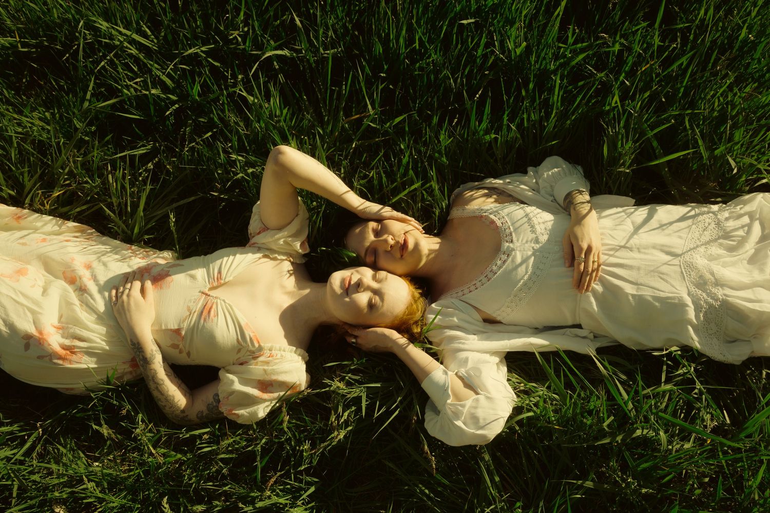 Two women in floral dresses lying side by side on green grass, eyes closed, in soft golden light.