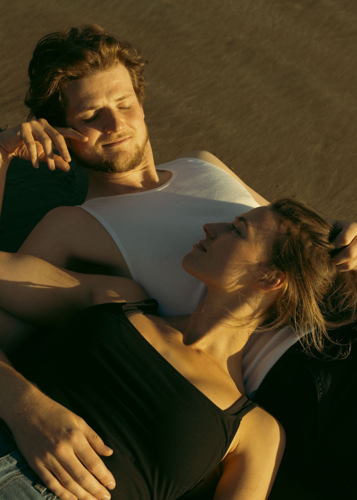 Couple relaxing outdoors in golden sunlight, woman resting on mans chest, both with eyes closed in peaceful embrace.