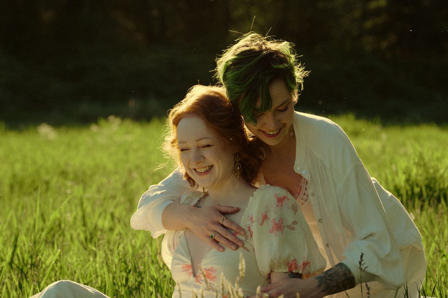 Two women laugh joyfully together while sitting in a sunlit green meadow on a warm day.