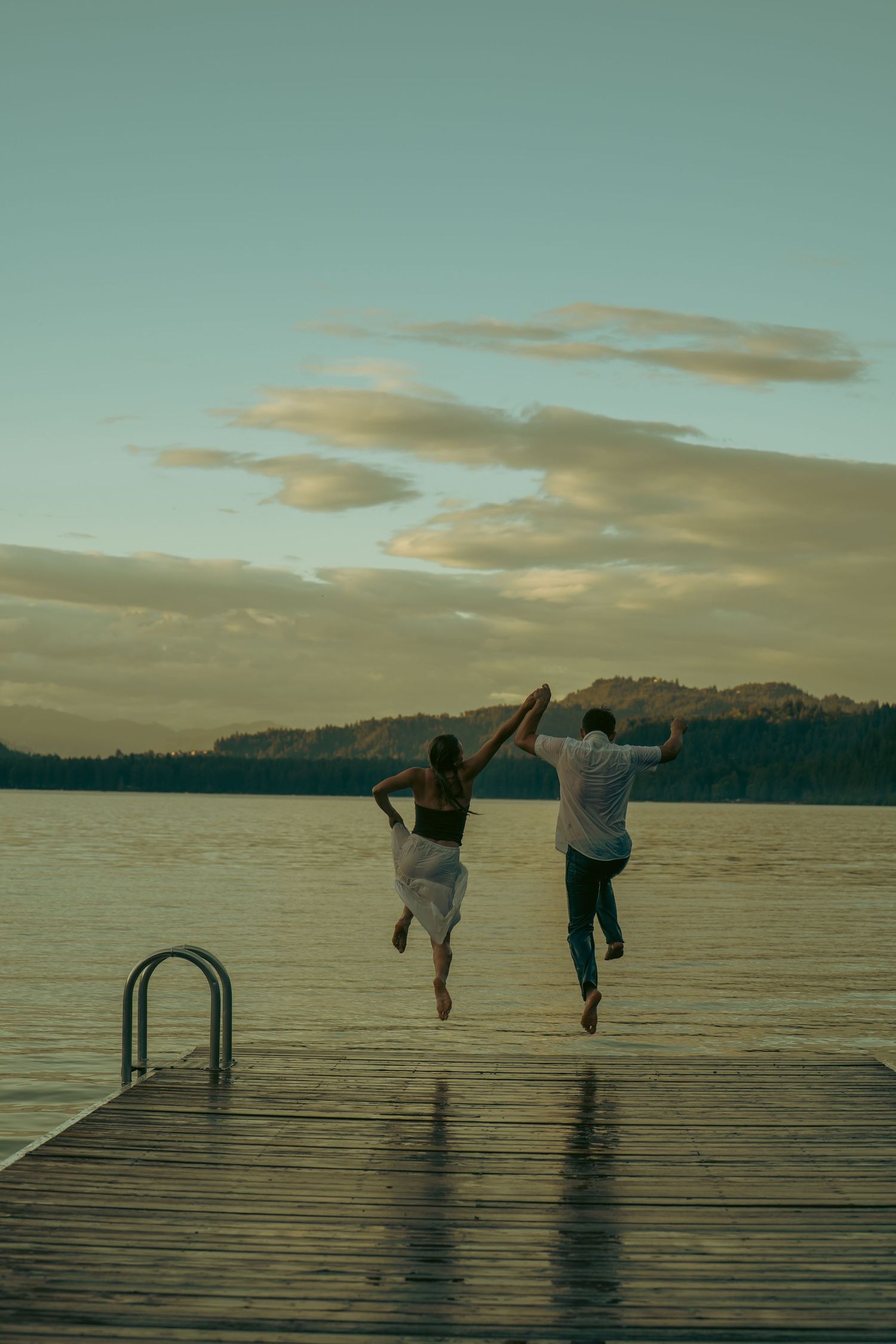 Couple jumping off a wooden dock into a lake at sunset with cloudy sky and forested hills in background.