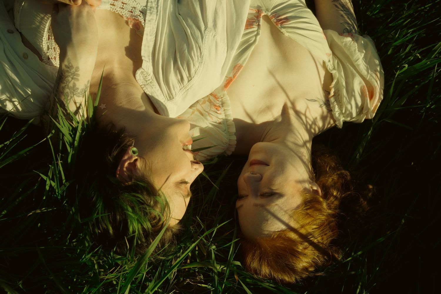 Two people lying in green grass, heads together, bathed in dappled sunlight.