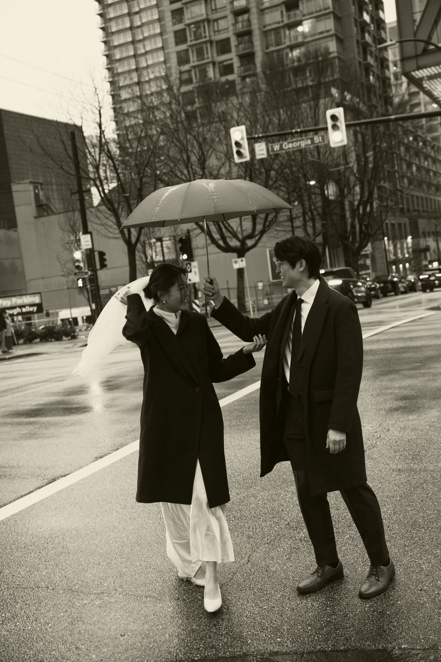 Black and white photo of a couple sharing an umbrella on a rainy city street, dressed in elegant coats.