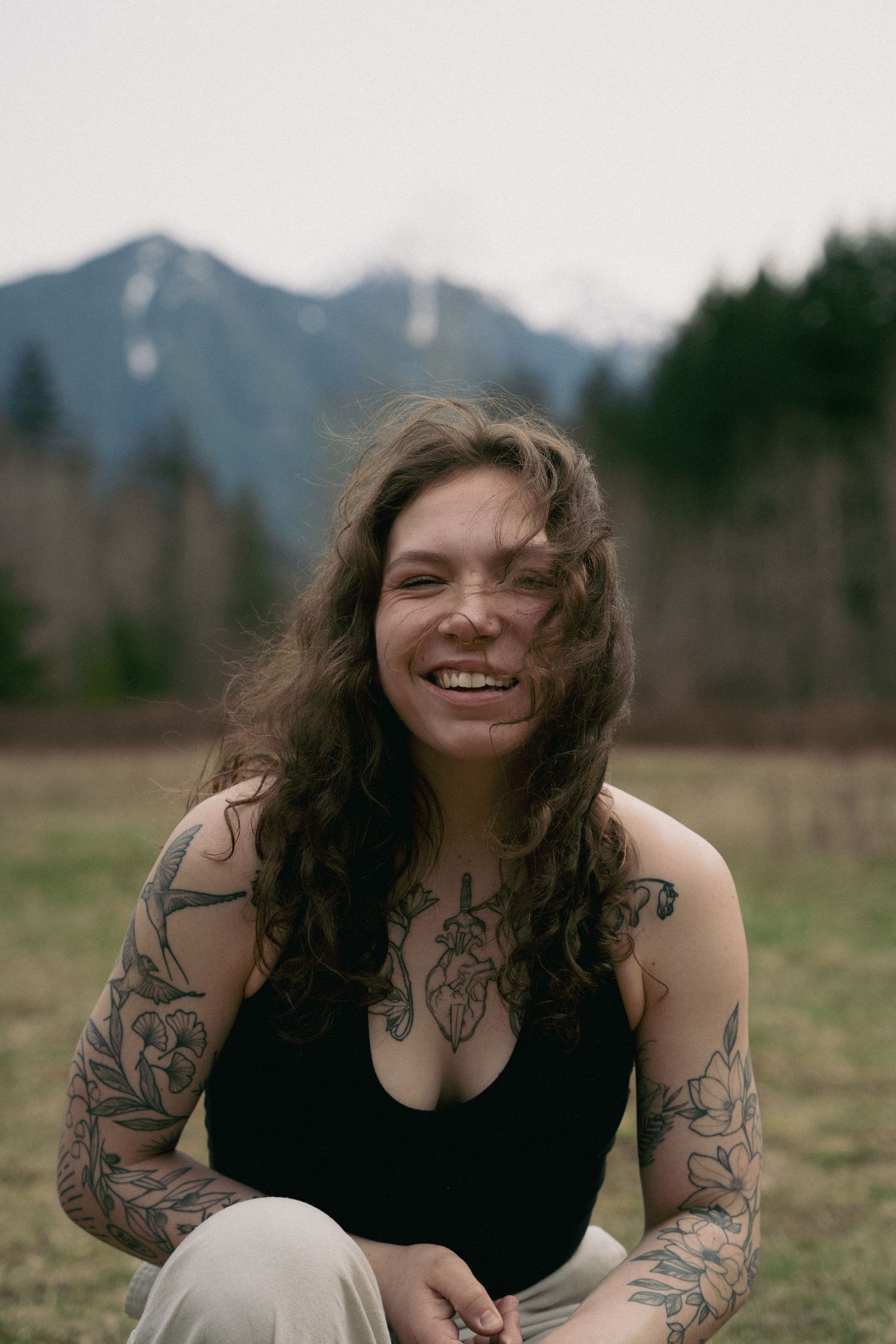 A person with dark curly hair and tattoos sits outdoors in a grassy field with mountains visible in the background.