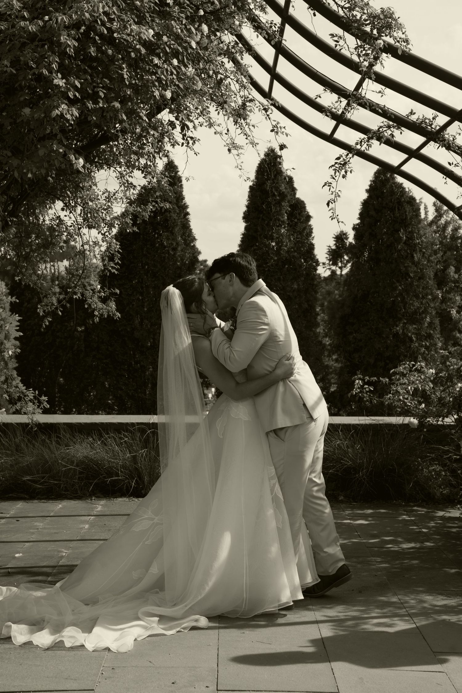 Black and white photo of bride and groom sharing first kiss under garden pergola.